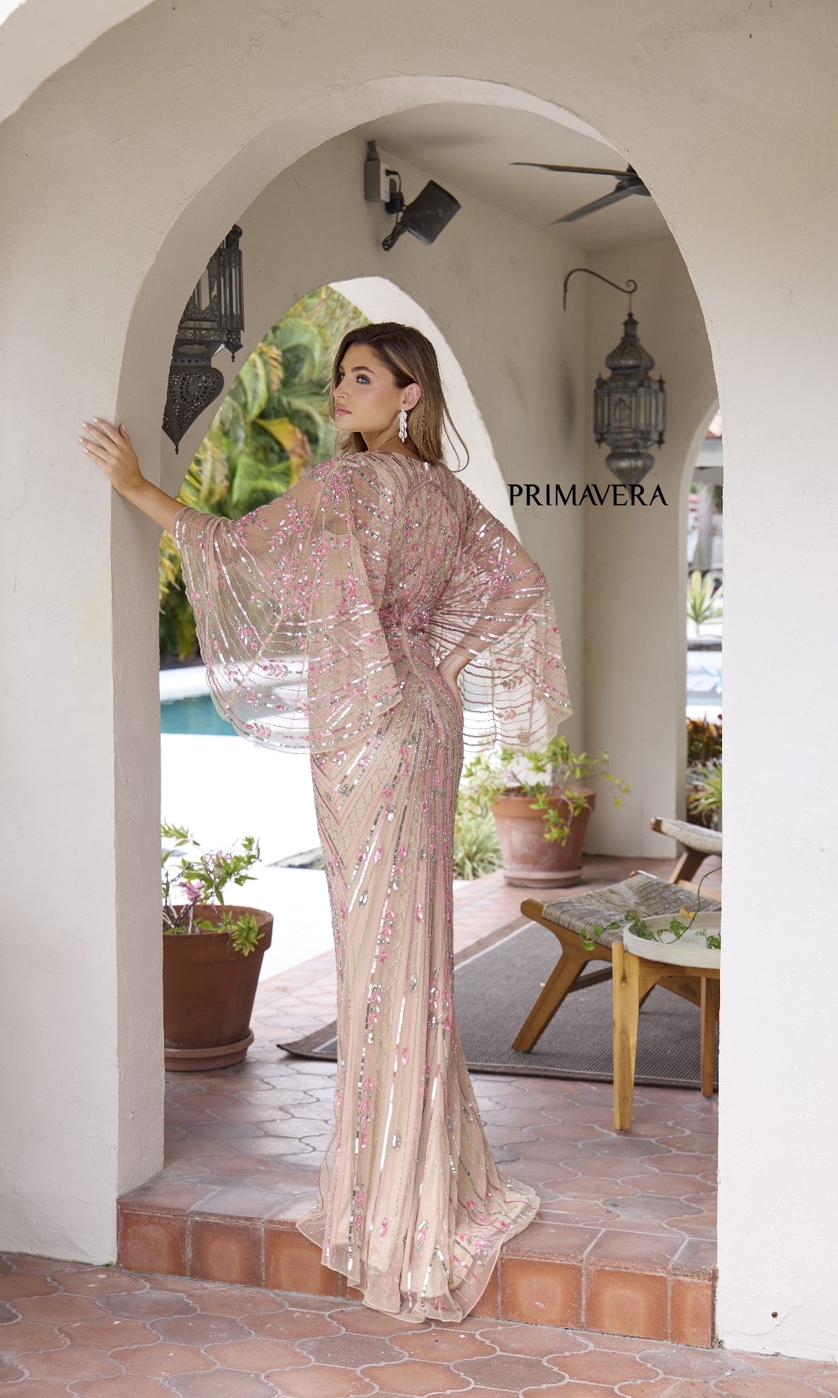 A woman, seen from behind, stands outdoors under an archway in front of plants and a pool, wearing the Primavera 12189 Long Formal Dress—a sheer beaded gown with a v-neckline. 