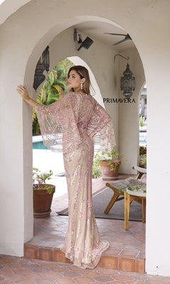 A woman, seen from behind, stands outdoors under an archway in front of plants and a pool, wearing the Primavera 12189 Long Formal Dress—a sheer beaded gown with a v-neckline. "PRIMAVERA" is visible on the wall.