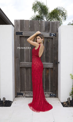 A woman poses in front of a wooden gate wearing the Primavera 12192—a long, fitted red formal dress with a sleeveless design, open back, and v-neckline.