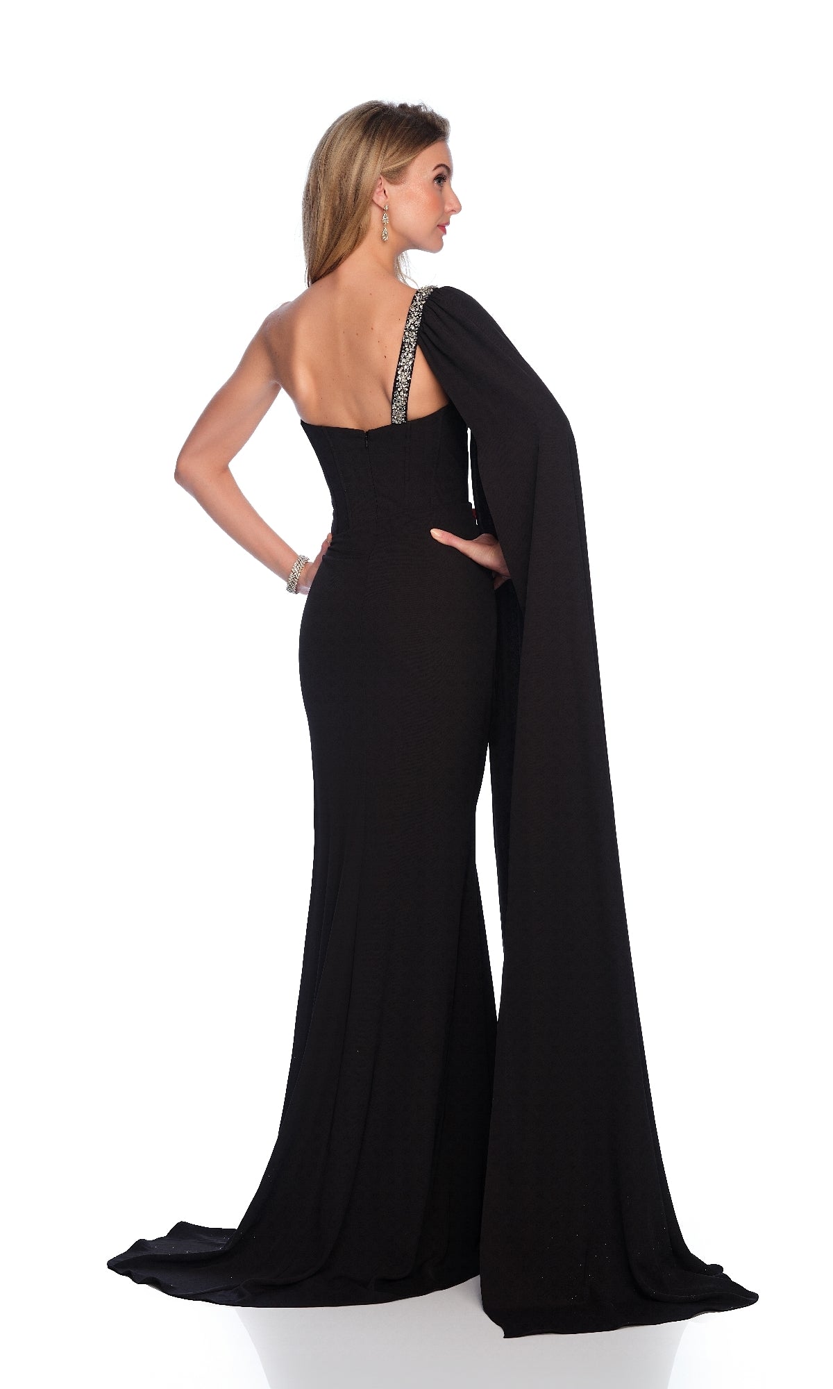 A woman stands facing away against a white background, wearing the Long Black Prom Dress: Dave & Johnny 12232, featuring a one-shoulder design, draped cape detail, and elegant corset bodice.