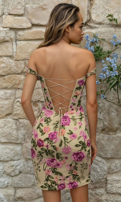 A woman stands before a stone wall in the Short Homecoming Dress 12370: an off-the-shoulder floral lace dress with corset bodice, lace-up back, and pink and purple flowers on a light background.
