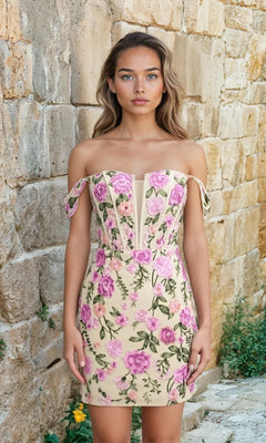 A woman stands against a stone wall wearing the Short Homecoming Dress: 12370—an off-the-shoulder, fitted beige dress with pink floral patterns and a delicate corset bodice.