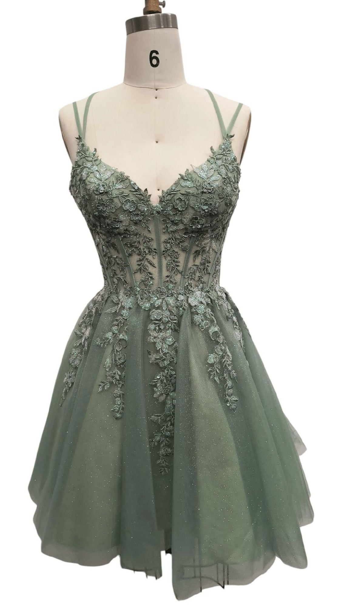 A mannequin showcases the Short Sage Green Hoco Dress: 12435, featuring sheer panels, floral lace appliqué, thin straps, and a fitted bodice—a showstopping choice for your next party.