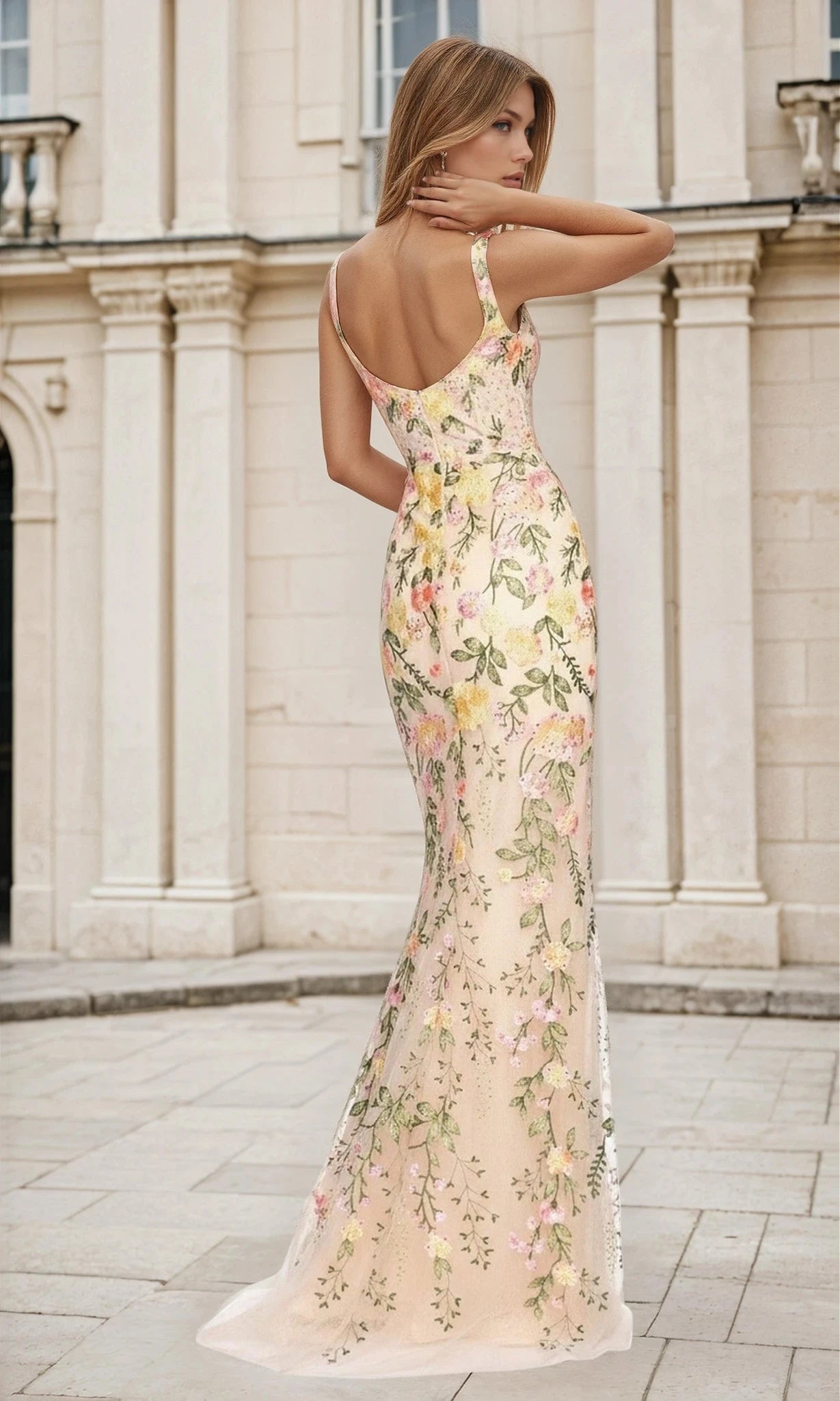 A woman in the Open-Back Floral-Print Nude Long Prom Dress L487 stands outdoors, facing away from the camera before a light-colored stone building.