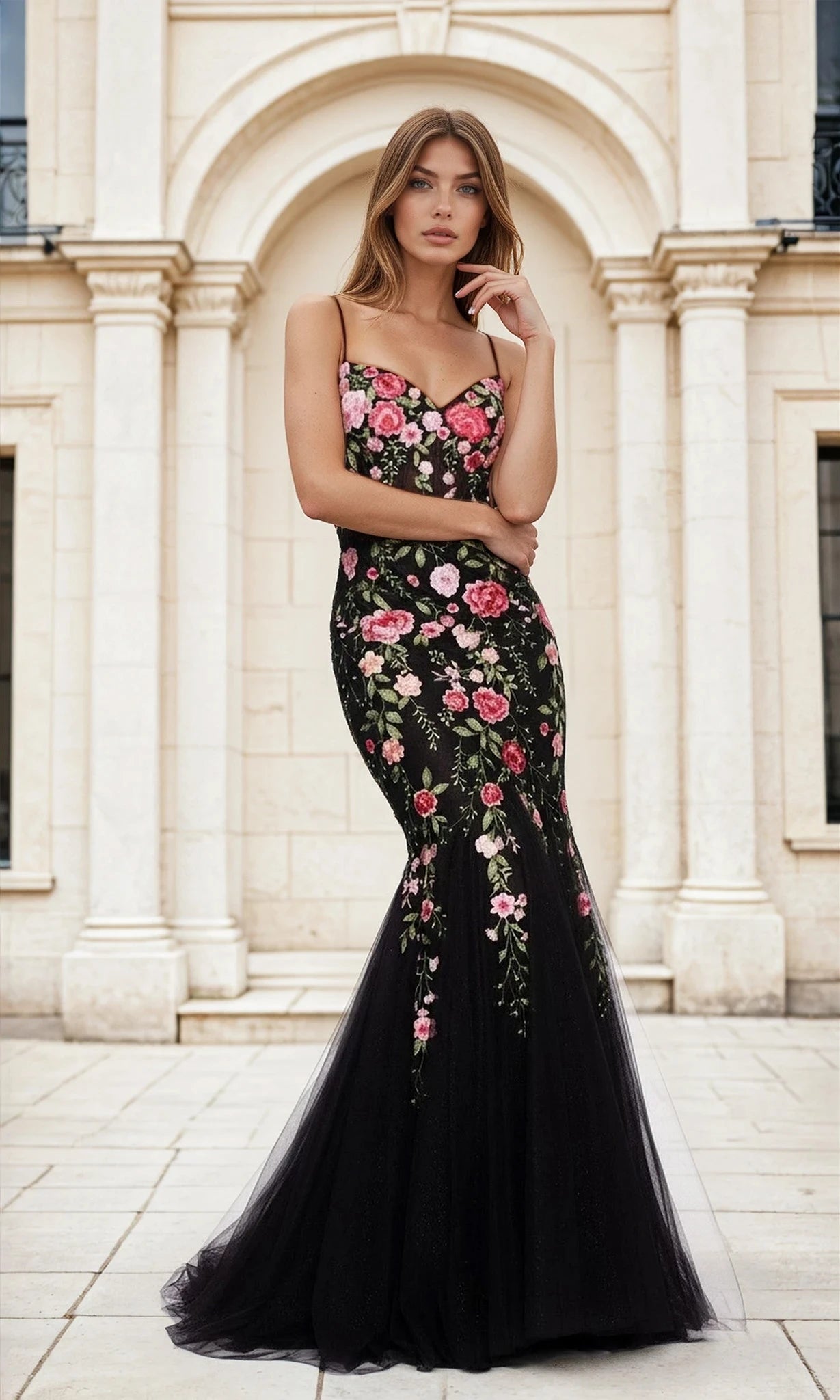 A woman stands outdoors before a stone building, wearing the Floral-Print Black Glitter Mermaid Prom Dress L488 featuring pink floral accents and a flared tulle skirt.