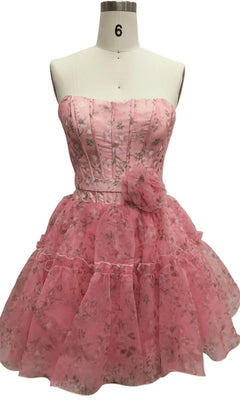The Strapless Short Pink Hoco Dress: 12603 features a floral print, corset-style bodice, layered ruffled skirt, and waist flower detail. Displayed on a dress form, it's the perfect pink party mini dress.