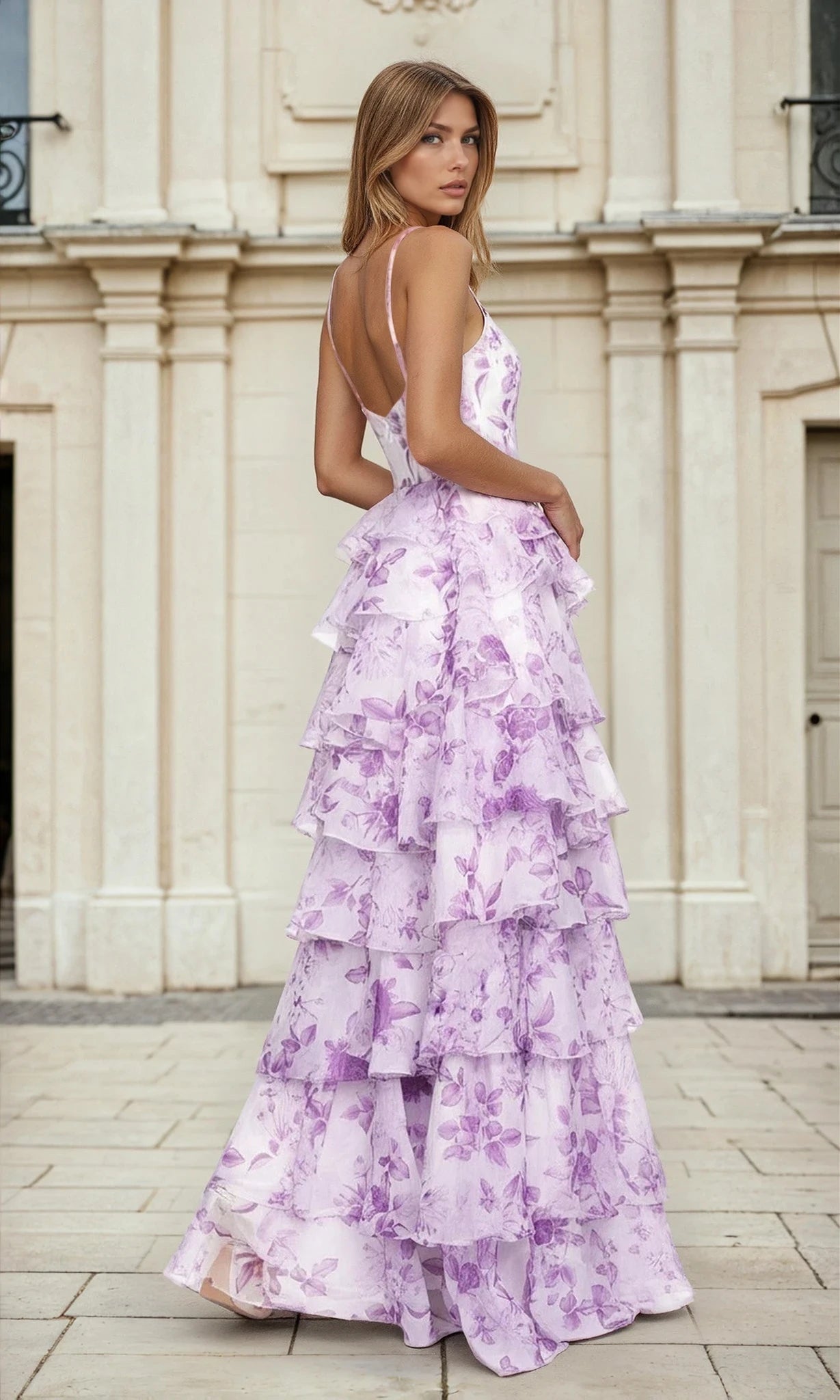 A woman stands outdoors in the Purple Floral A-Line Tiered Prom Dress L691, featuring a sleeveless design, ruffled A-line skirt, and boned bodice as she looks over her shoulder.