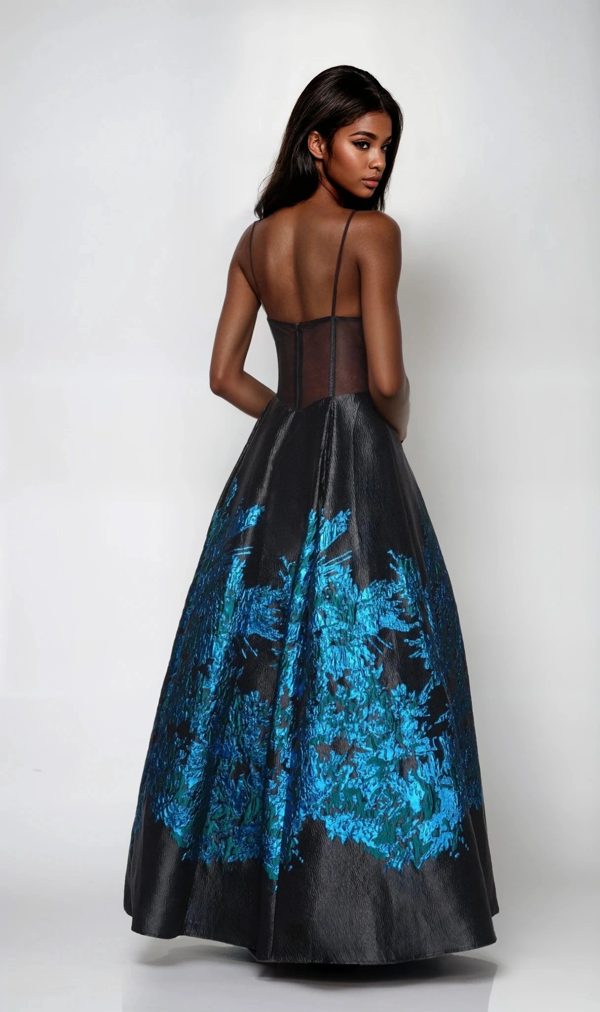 A woman faces away in a Dave and Johnny 12702 long prom dress featuring a black corset bodice, sheer back, and blue floral patterns on the full skirt, set against a white background.