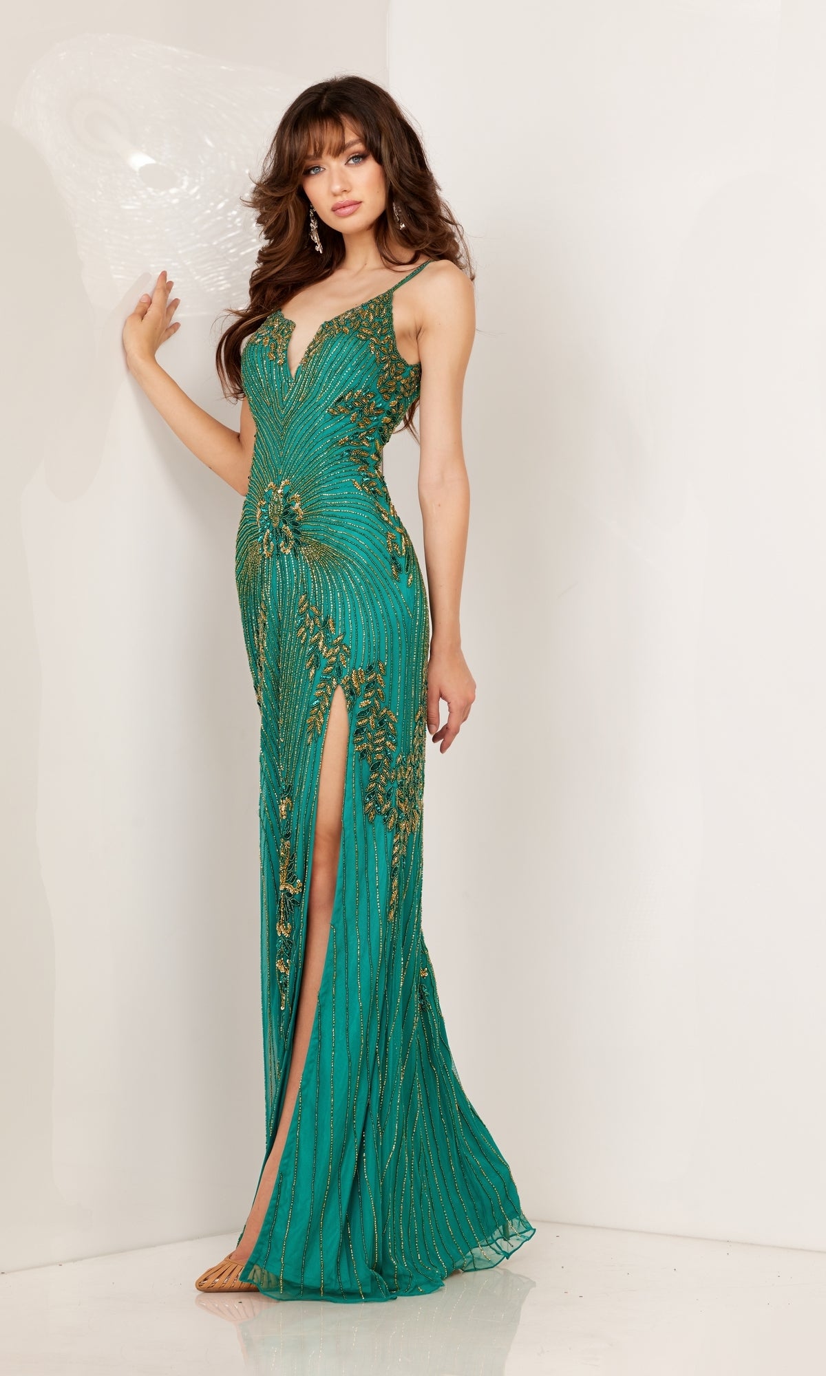 A woman wears the Aleta Long Formal Prom Dress 1275, a fitted green gown with gold embellishments, beading, and a thigh-high slit, standing against a white wall.
