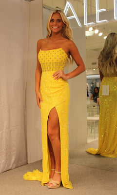 A woman stands indoors in the Aleta Strapless Beaded Designer Prom Dress 1278, a yellow sparkly gown with a thigh-high slit. She pairs it with neutral heels and lets her long wavy hair flow, while a mirror shows off the dress's glamorous back.