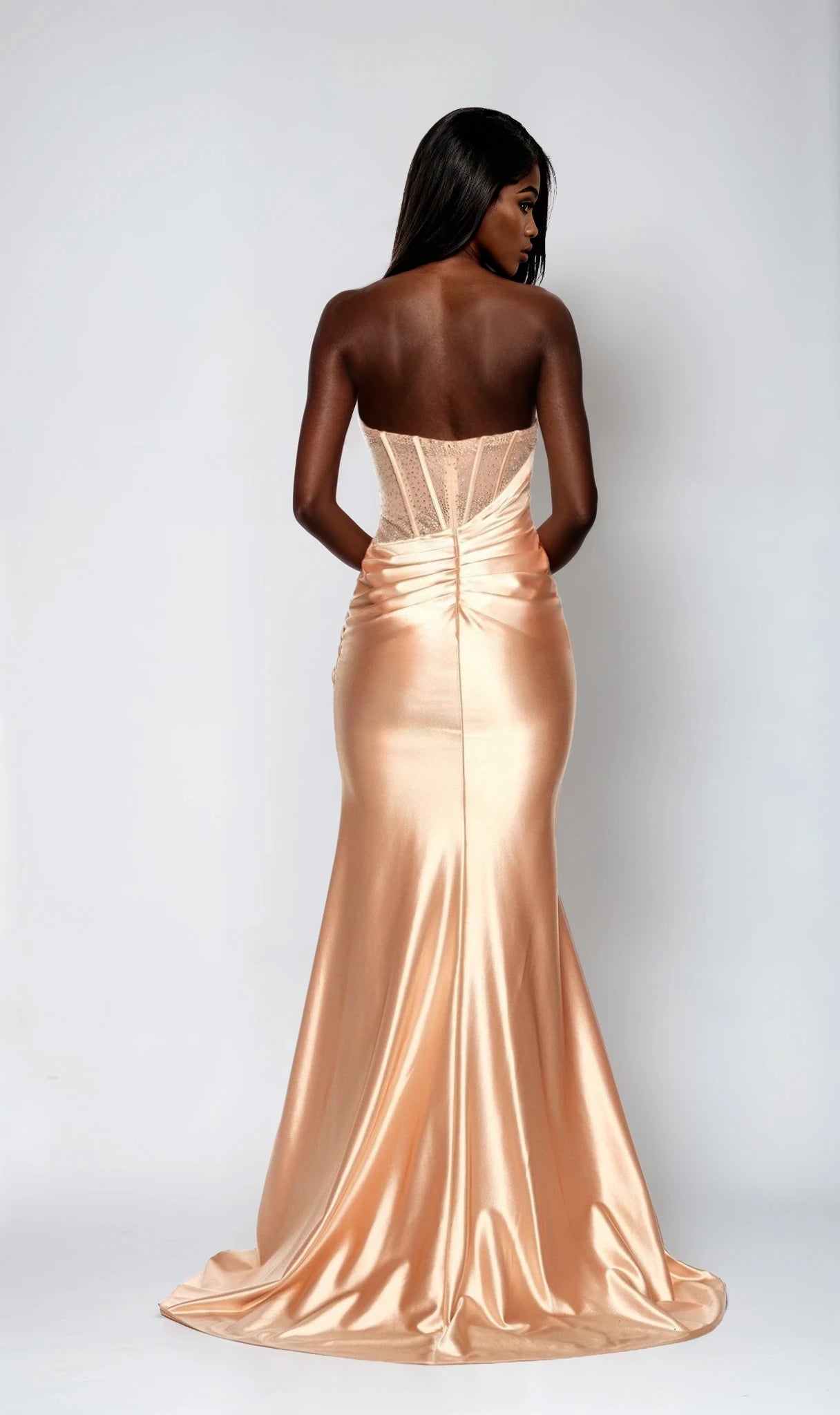 A woman faces away in the Dave and Johnny 12856 long prom dress, a strapless, fitted champagne satin gown with floor-length design and ruched back detail, set against a plain light background.