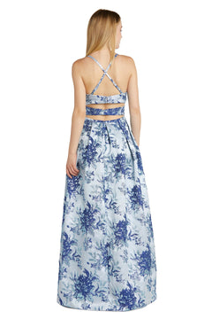 A woman with long blonde hair is seen from behind wearing the Morgan 12945 Blue Floral A-Line Prom Dress, which features a floor-length skirt and a strappy cut-out back.