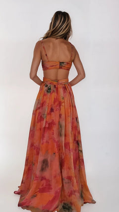 A woman stands facing away, wearing the Dave and Johnny 12955 long prom dress. This orange and pink patterned chiffon gown features thin straps, an open back, a flowy A-line skirt, and drapes gracefully against a plain background.