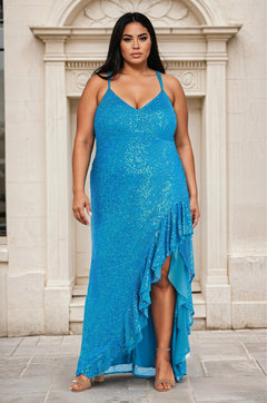 A woman in the Long Plus-Size Sequin Prom Dress Morgan 13029WM with an empire waist and ruffled asymmetrical hem stands outside before an ornate stone archway.