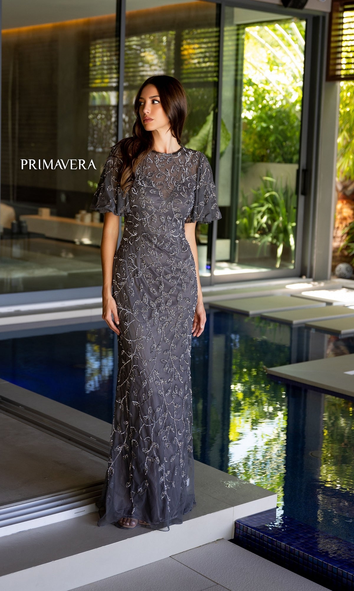 A woman in a fitted Long Prom Dress 13111 by Primavera stands by an indoor pool with glass walls and lush greenery outside. 