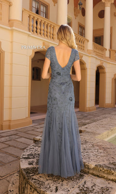 A woman wearing the Long Prom Dress 13112 by Primavera, a light blue floral beaded mermaid gown with short sleeves and an open back, stands outdoors before a beige building with arches and columns.