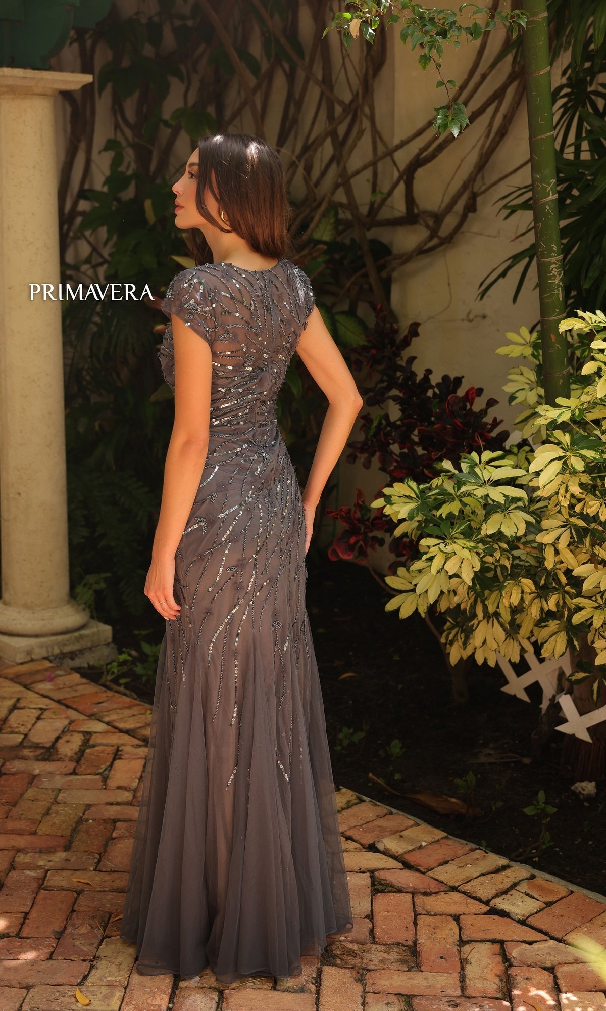 A woman wears the Long Prom Dress 13114 by Primavera, a gray, floor-length, short-sleeved fitted formal gown with sequin detailing, while standing on a brick patio among green and yellow foliage.