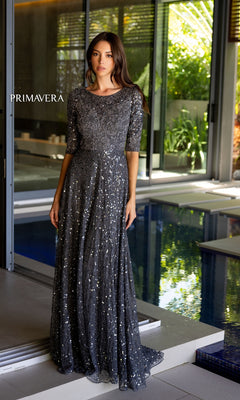 A woman stands indoors by a pool wearing the Long Prom Dress 13115 by Primavera, a floor-length silver-gray A-line beaded formal dress with half sleeves. The word "PRIMAVERA" appears on the left side of the image.