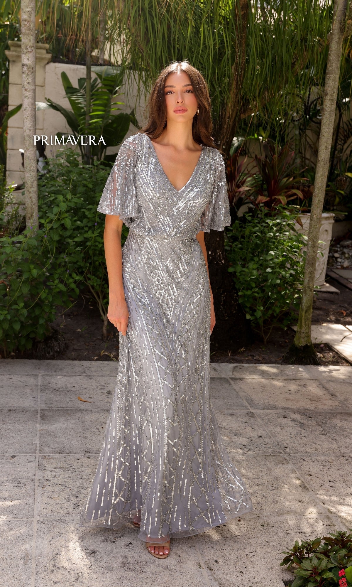 A woman stands outdoors among lush greenery and stone tiles, wearing the Primavera 13119 Sleeved Long Sequin Formal Dress, a silver floor-length gown with short sleeves and intricate sequin patterns. 