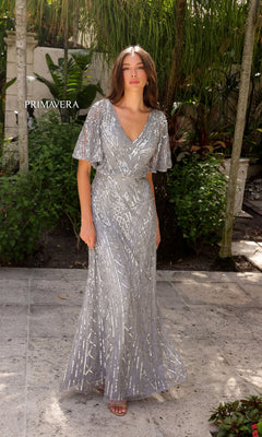 A woman stands outdoors among lush greenery and stone tiles, wearing the Primavera 13119 Sleeved Long Sequin Formal Dress, a silver floor-length gown with short sleeves and intricate sequin patterns. "PRIMAVERA" is displayed in the background.