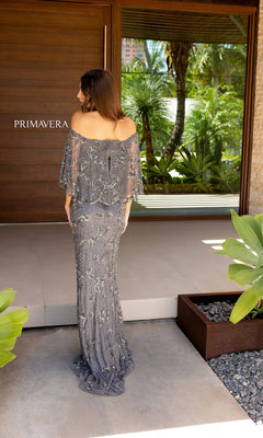 A woman in the Caped Long Beaded Prom Dress: Primavera 13123 stands on a modern patio facing away, with tropical plants and a wooden door in the background.