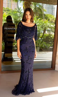A woman stands indoors wearing the Caped Long Beaded Prom Dress: Primavera 13123, a navy blue evening gown. Palm trees and buildings are visible through a glass door behind her.