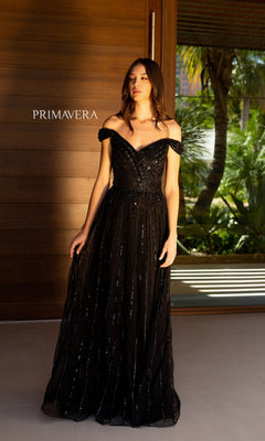 A woman in a black Long Prom Dress 13125 by Primavera with sequins stands before a wooden wall featuring the "PRIMAVERA" logo, with a glass door and greenery visible outside.