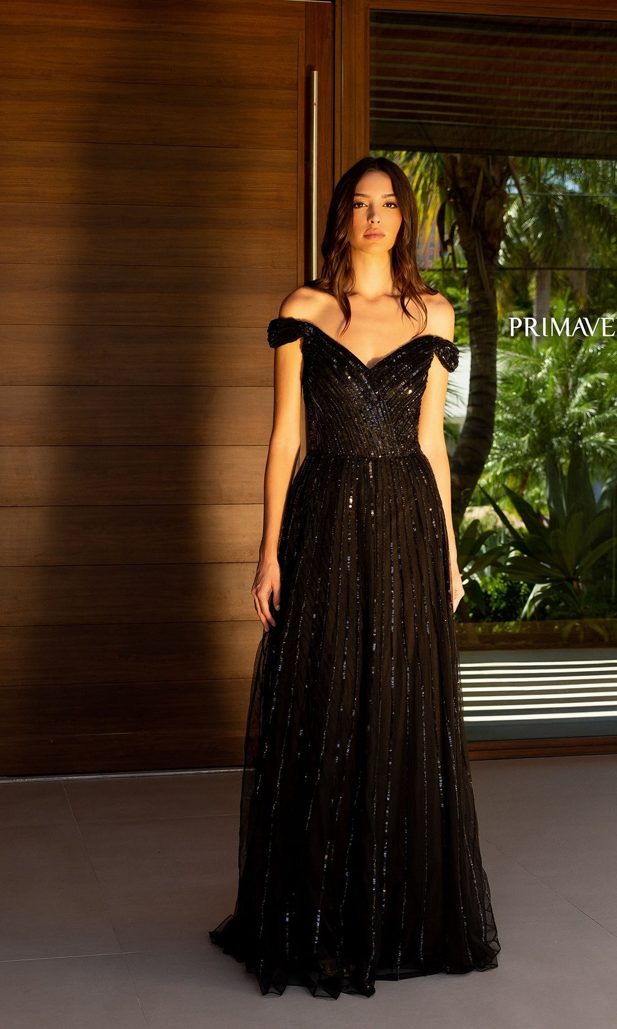 A woman stands indoors wearing the black Long Prom Dress 13125 by Primavera, featuring sequin embellishments. Through the window behind her, tropical scenery is visible.