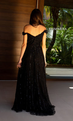 A woman in a black, off-the-shoulder Long Prom Dress 13125 by Primavera, featuring sequin details, stands indoors with her back turned. The word "PRIMAVERA" is displayed beside her.