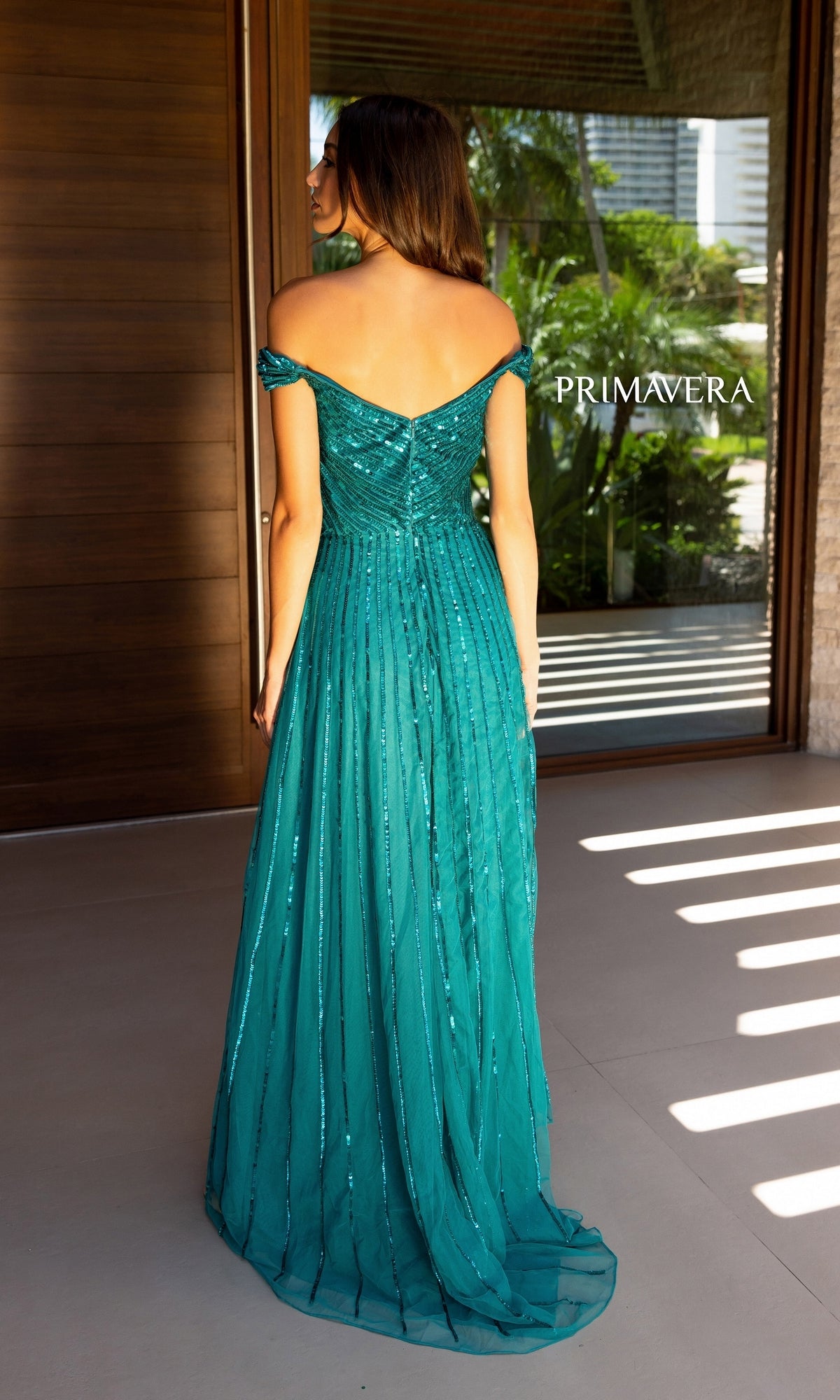 A woman, seen from behind near a modern building and greenery, wears the shimmering Long Prom Dress 13125 by Primavera. The word 