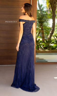 A woman faces away indoors near a wooden door, wearing the Primavera 13126 off-shoulder beaded prom dress in navy with subtle sparkly embellishments. Greenery can be seen outside.