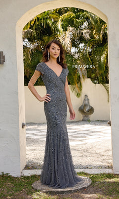 A woman stands under an outdoor archway with greenery, wearing the Primavera 13133—a long formal dress featuring floral beading, cap sleeves, and a v-neckline.
