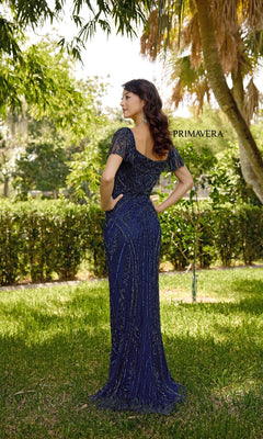 A woman stands outdoors in the navy blue Primavera 13134 long formal dress, featuring a sweetheart neckline, delicate beading, short sleeves, and an open back, with lush trees and greenery behind her.
