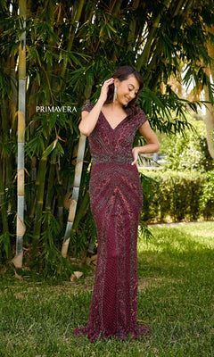 A woman stands on grass near bamboo, smiling and touching her earring while wearing the Primavera 13135: a deep red, beaded long formal dress with a V-neck and fitted silhouette. "PRIMAVERA" appears on the left side of the image.