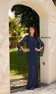 A woman stands beneath a stone archway in the Primavera 13136 long formal dress, featuring navy blue embellishments and sheer bell sleeves. "PRIMAVERA" appears on the wall, with trees and grass in the background.
