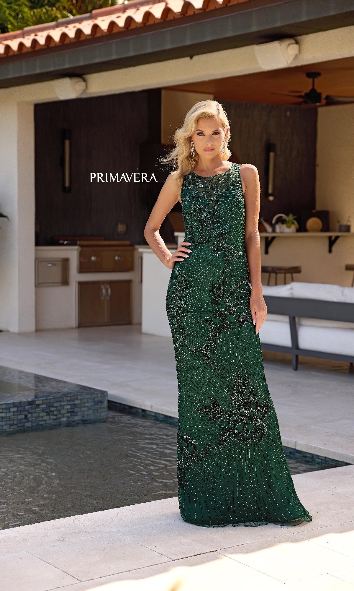 A woman wearing the Primavera 13140 sleeveless long formal dress with an illusion-sweetheart neckline stands by a pool on an outdoor patio. The word 