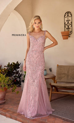 A woman in the Primavera 13143 long formal dress, featuring a light pink hue, sweetheart neckline, and sparkly embellishments, stands on a tiled patio near outdoor furniture and potted plants under an archway.