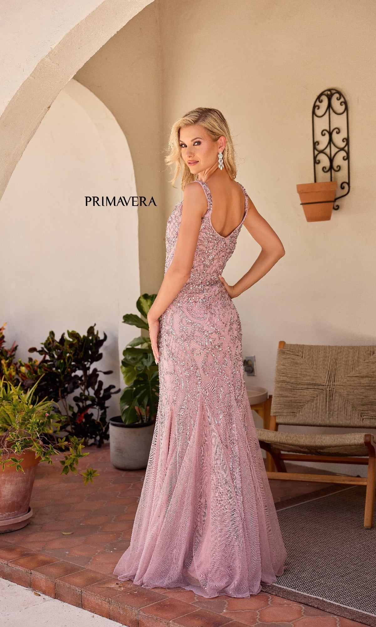 A woman in the Primavera 13143 long formal dress—a sleeveless, floor-length pink sequin gown with a sweetheart neckline—stands on a patio, posing near plants and outdoor furniture as she looks over her shoulder.