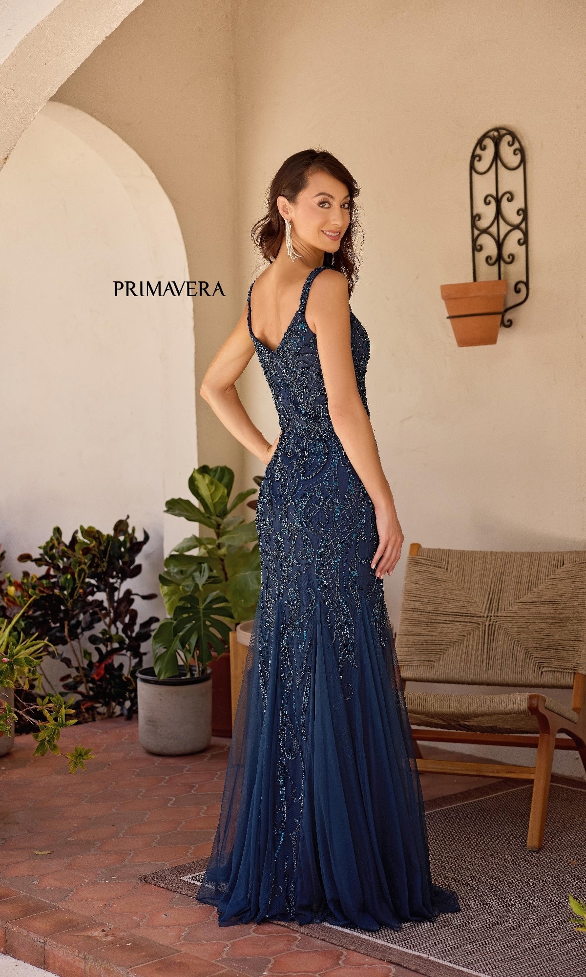 A woman in a navy blue Primavera 13143 long formal dress with sparkly embellishments stands on a patio near potted plants and a wicker chair, facing away from the camera.
