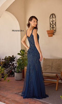 A woman in a navy blue Primavera 13143 long formal dress with sparkly embellishments stands on a patio near potted plants and a wicker chair, facing away from the camera.