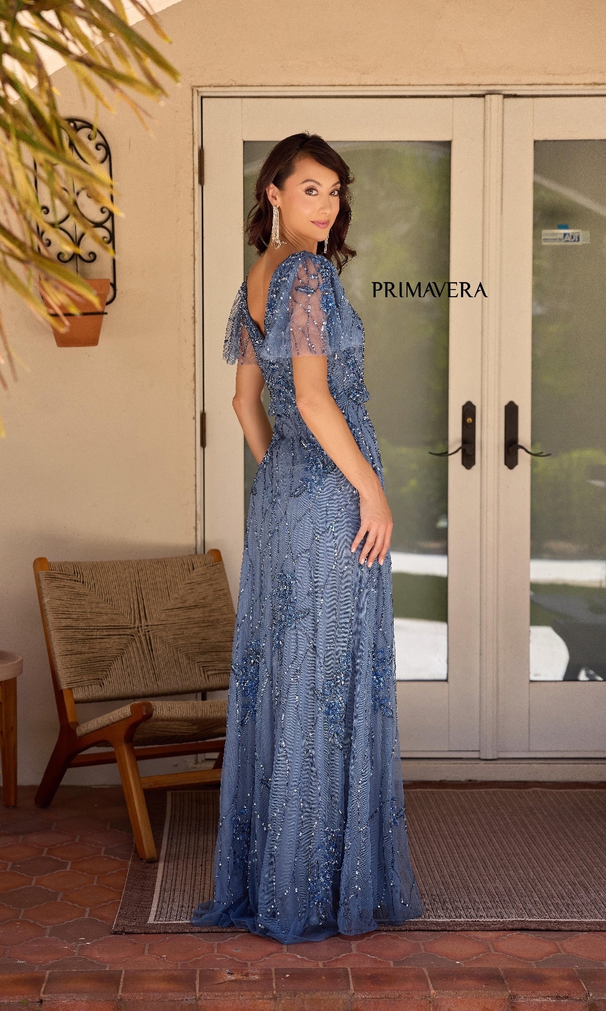 A woman in a blue, beaded Long Formal Dress: Primavera 13144 stands on a porch facing a glass door labeled 