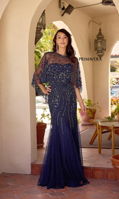 A woman stands under an archway wearing the Primavera 13145: a navy blue, beaded, strapless long formal dress with a mermaid skirt and a sheer, embellished removable capelet.