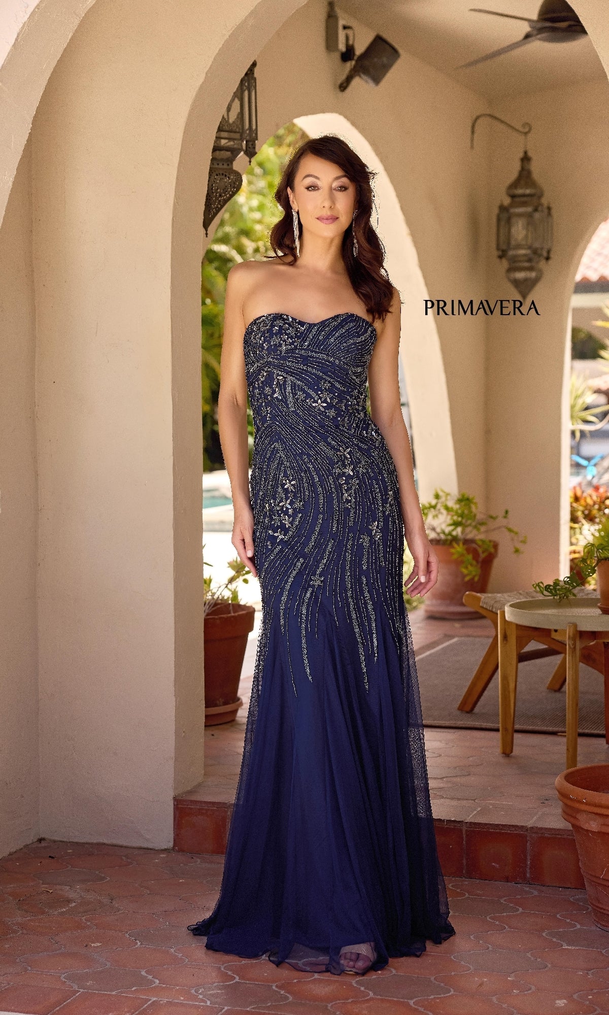 A woman stands under an archway wearing the Sheer Capelet Long Formal Dress: Primavera 13145, a floor-length navy blue gown with shimmering beadwork and a mermaid skirt.