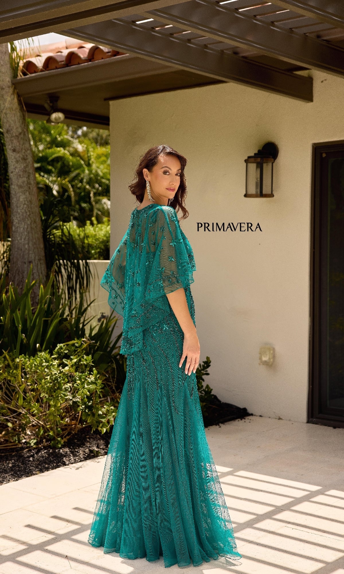 A woman stands outdoors on a patio near greenery and a house wall with 