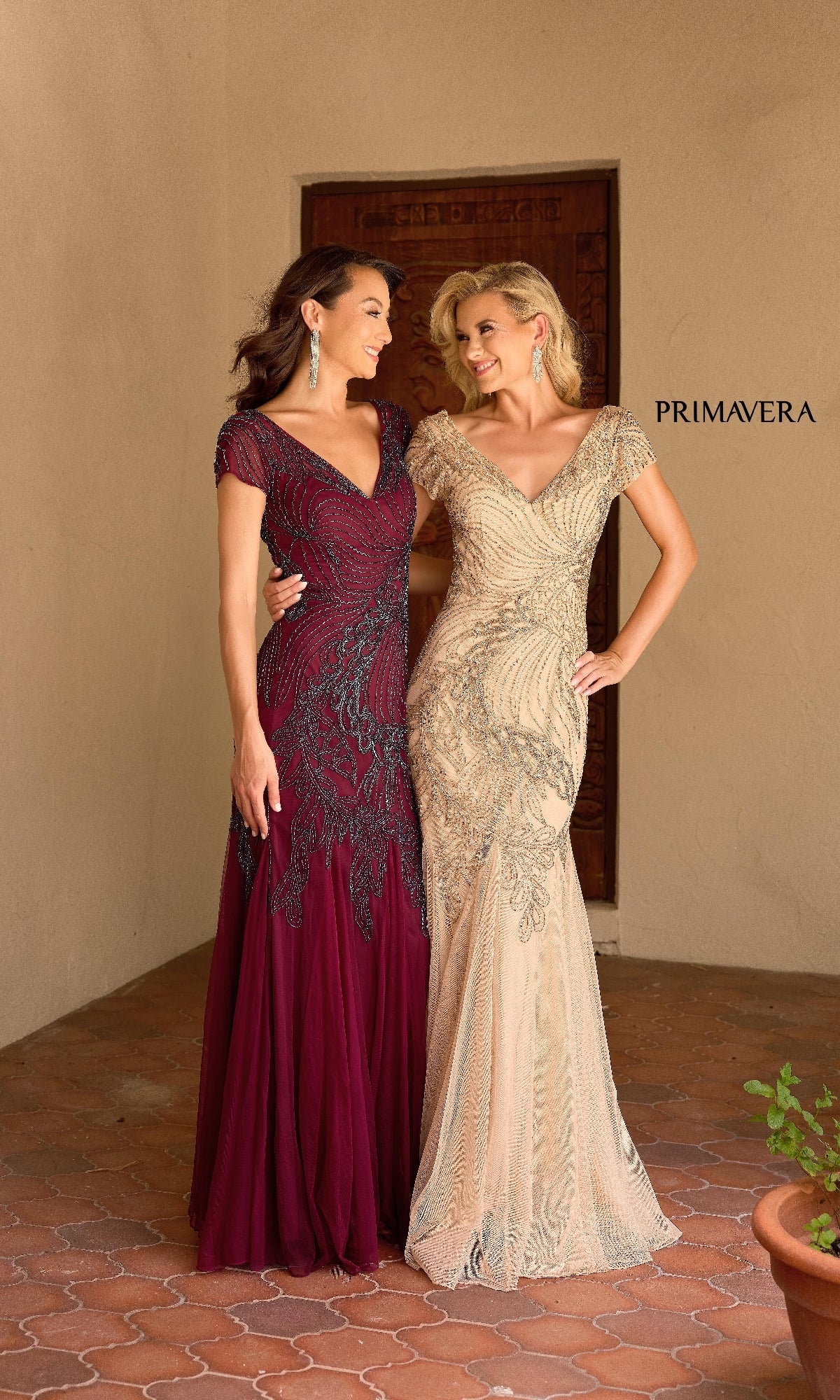 Two women in elegant floor-length gowns stand side by side, smiling in a tiled hallway. One wears the Primavera 13148 long formal dress; behind them, 