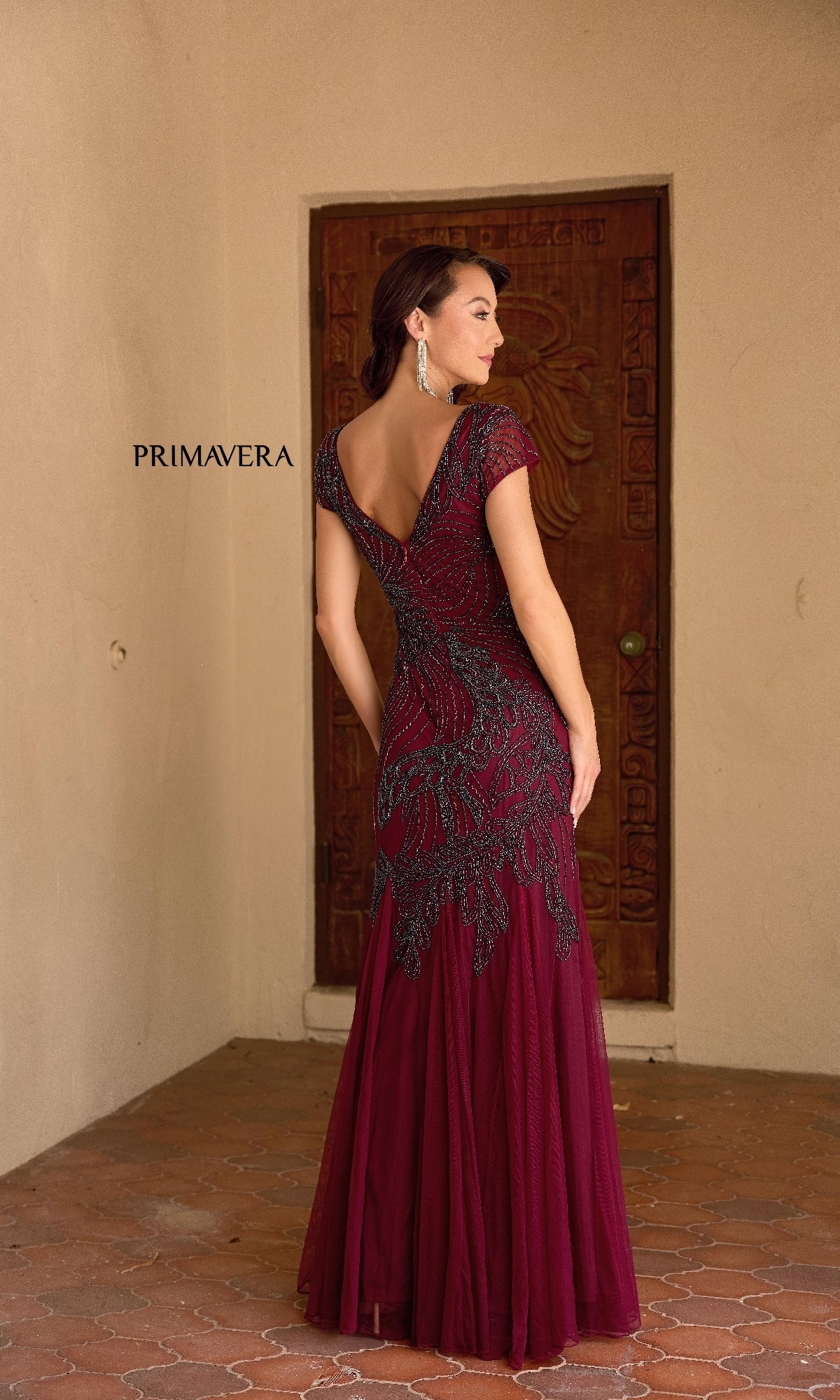 A woman wearing the Primavera 13148 long formal dress in burgundy and black with beaded embellishments stands with her back to the camera before a wooden door. 