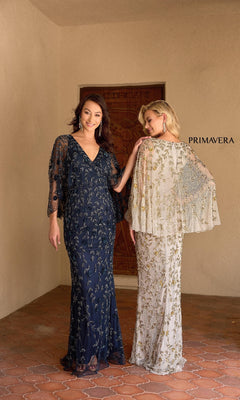 Two women wear Primavera 13149 long formal gowns; one faces front in a navy blue v-neck dress, the other shows the back of a cream gown. "PRIMAVERA" appears on the wall behind them.