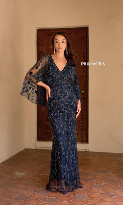 A woman stands in front of a wooden door wearing the Primavera 13149, a navy blue long formal dress featuring silver floral embroidery, beaded details, a v-neckline, and flared sleeves.