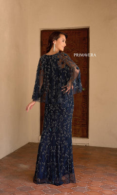 A woman poses indoors in the Primavera 13149, a navy blue, floor-length formal evening dress with beaded details, sheer sleeves, and a v-neckline. "PRIMAVERA" is displayed on the wall behind her.