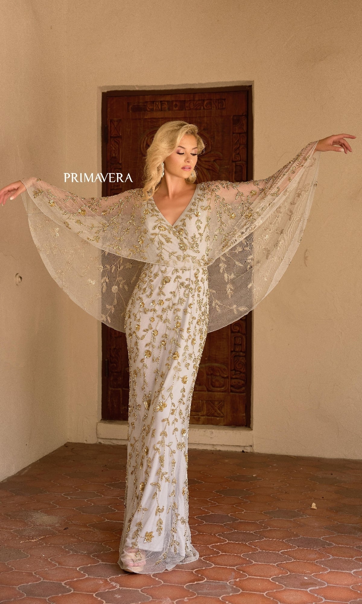 A woman poses with raised arms in front of a wooden door, showcasing the Primavera 13149 long formal dress—a white beaded gown with gold floral embroidery, v-neckline, and sheer wing-like sleeves.